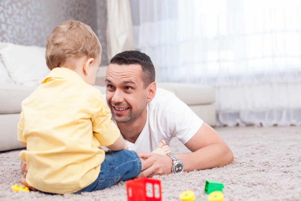 Attractive father is playing with his son and smiling. He is lying on flooring and looking at the boy with love. 
